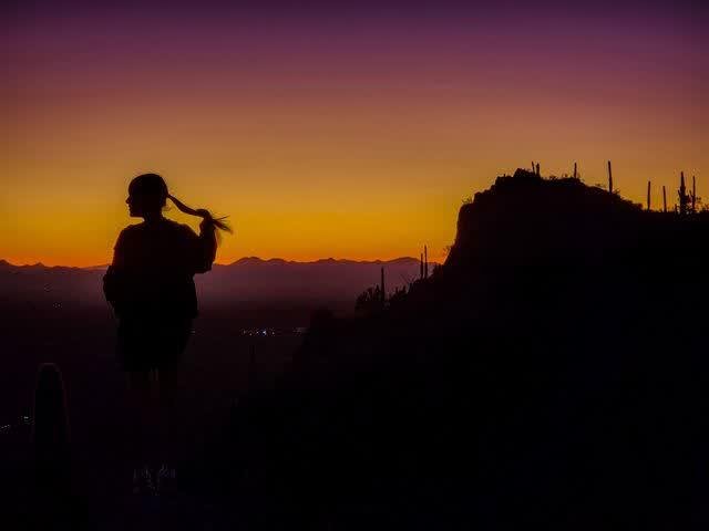 The silhouette of a woman with a ponytail holding her hair, standing against a vivid orange-to-purple sunset over desert mountains and saguaros.