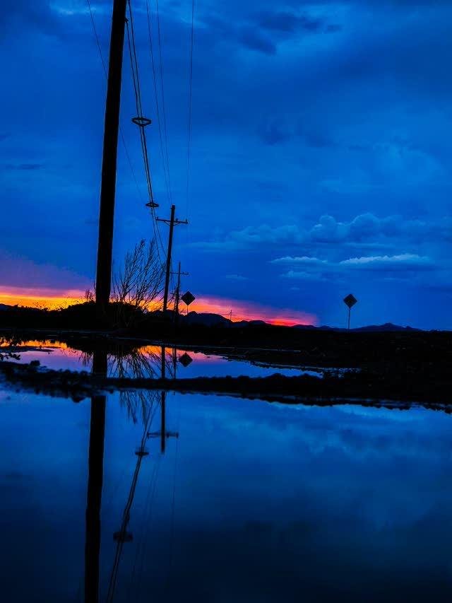 Power lines and utility poles silhouetted against a deep blue dusk sky, a band of orange-pink sunset on the horizon reflected in still water below.