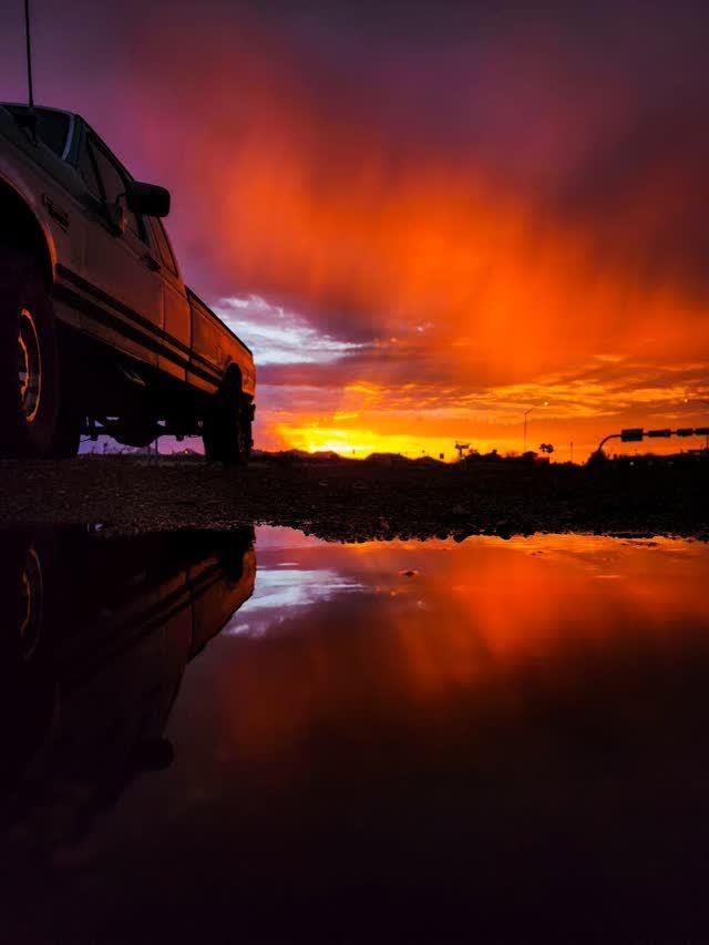 A pickup truck silhouetted against a blazing orange-red sunset, the truck and sky reflected in a puddle in the foreground.