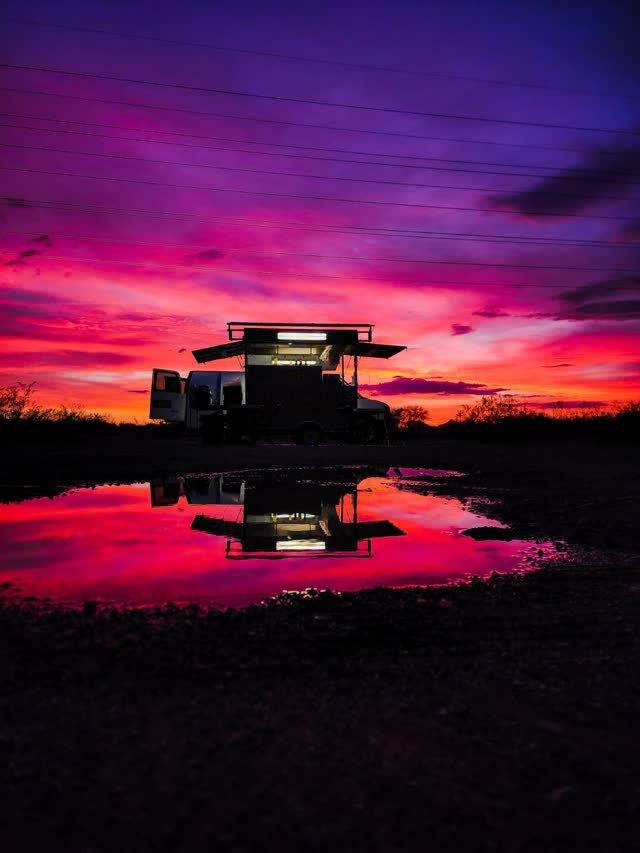 A food truck with its serving hatch open and lights on, silhouetted against a vivid pink-purple-orange sunset, reflected in a puddle below.