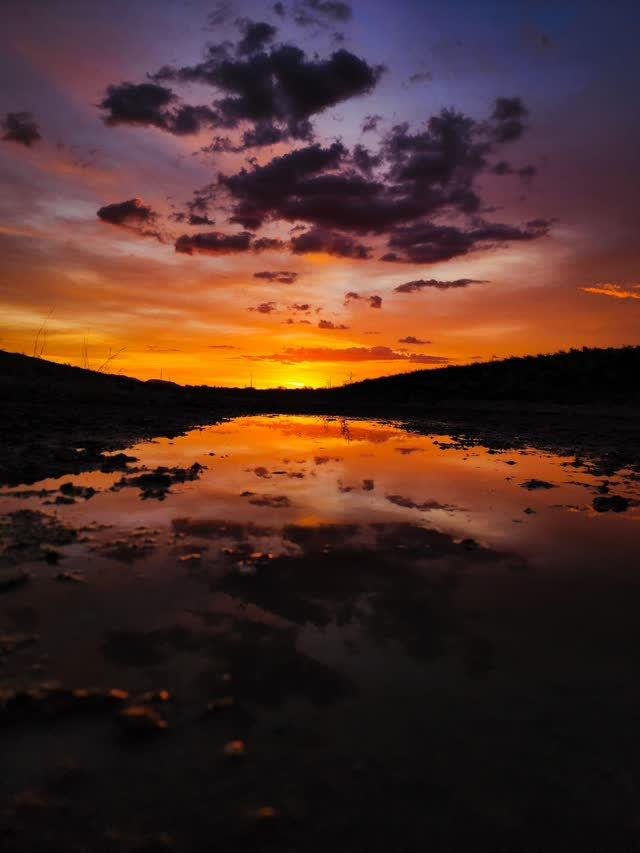 A blazing orange-red sunset with dark dramatic clouds mirrored in a pool of water in a desert wash, dark hills on the horizon.
